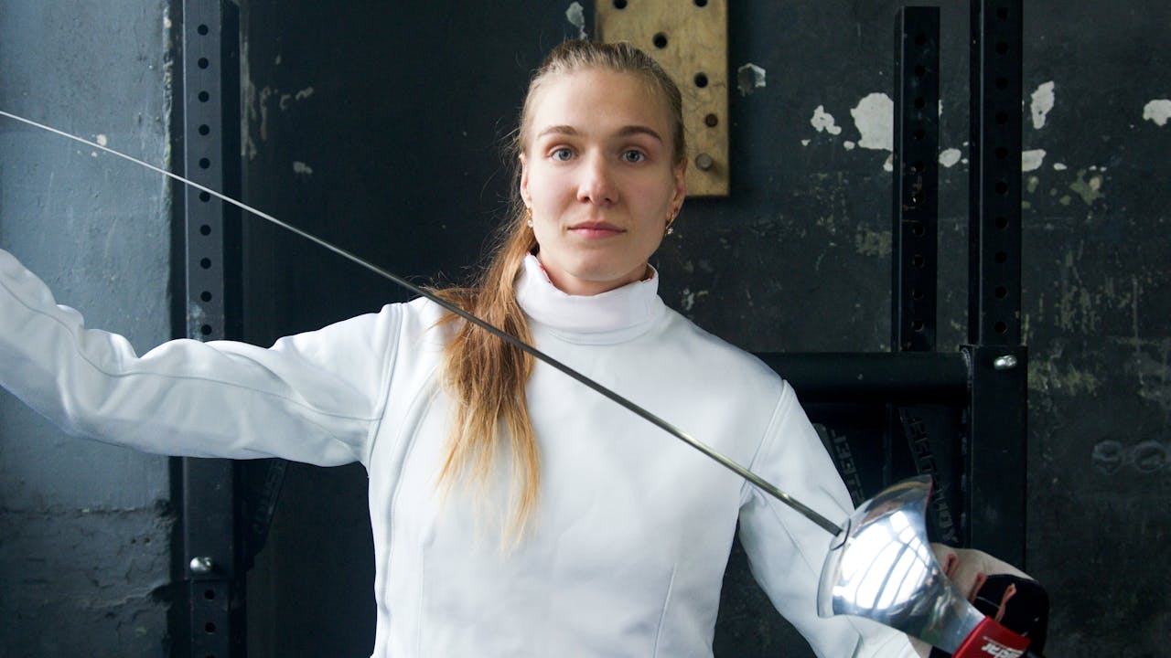 A woman in fencing attire holds a sword confidently in an indoor setting, ready for sport.