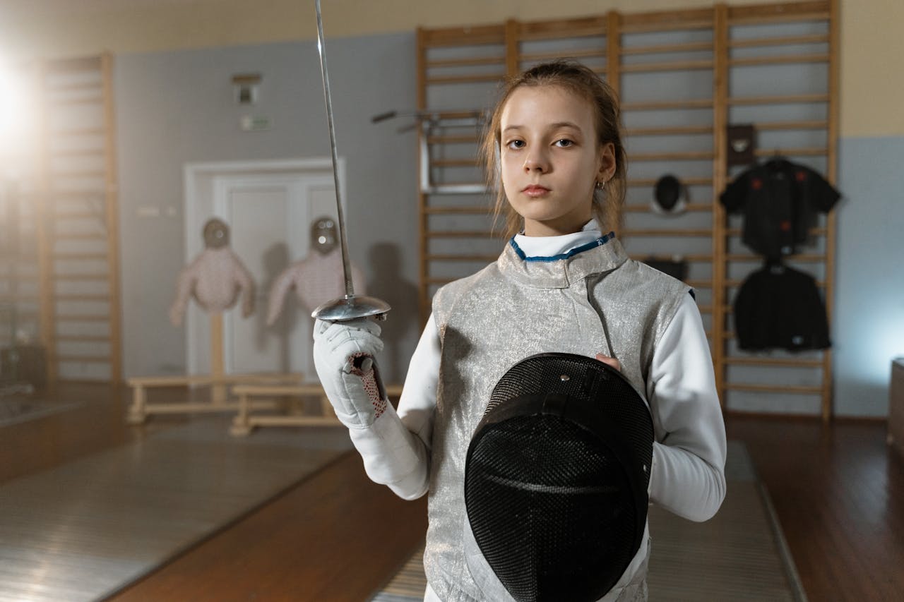 A young female fencer in full gear holding a foil and helmet during indoor training.